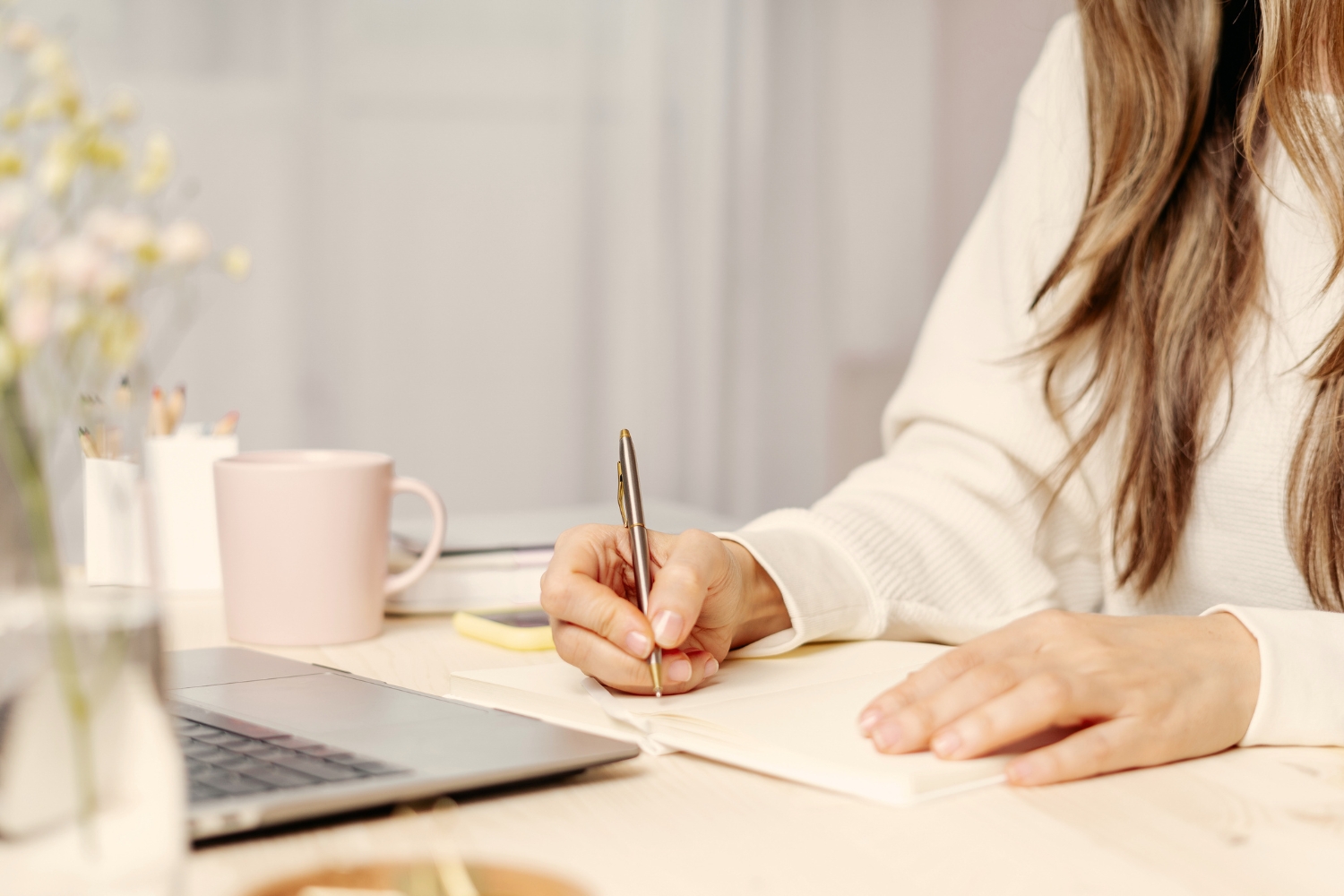 woman with long hair writing on notebook next to laptop and mug